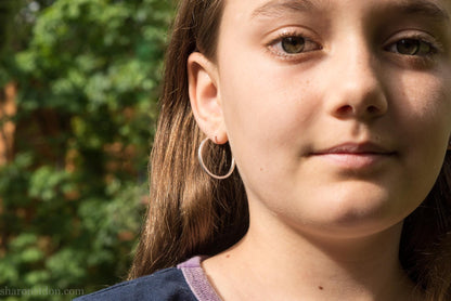 Hammered solid silver hoop earrings, handmade in the USA by Sharon Saint Don. Earrings are worn by a female child who is about 9 years old with fair skin and short brown hair. 25mm diameter, 2mm wide, 1.5mm thick. solid silver posts and backs. Lightweight and comfortable for daily wear with a matte, brushed, shiny finish.