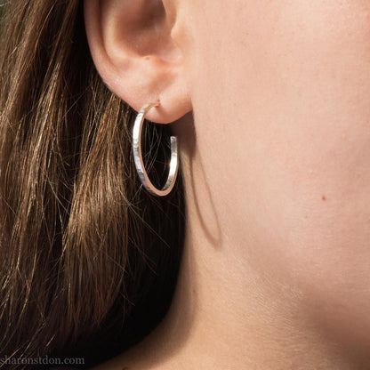 Hammered solid silver hoop earrings, handmade in the USA by Sharon Saint Don. Earrings are worn by a female child who is about 9 years old with fair skin and short brown hair. 25mm diameter, 2mm wide, 1.5mm thick. solid silver posts and backs. Lightweight and comfortable for daily wear with a matte, brushed, shiny finish.