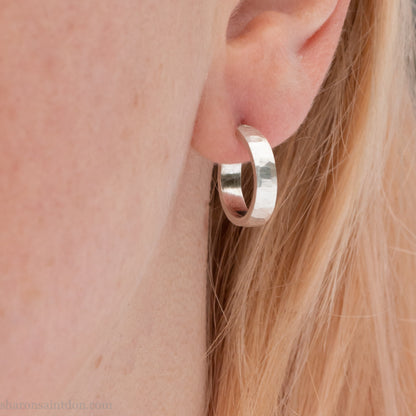A close-up view of a person wearing handcrafted solid silver hoop earrings, 14mm diameter x 3mm wide x 1.5mm thick with a hammered matte finish.