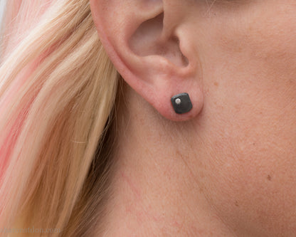 Person wearing a handcrafted, 7mm square, oxidized black sterling silver stud earring with a 2mm round moissanite gemstone set in the corner. 
