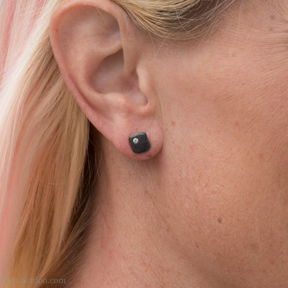 Person wearing a handcrafted, 7mm square, oxidized black sterling silver stud earring with a 2mm round moissanite gemstone set in the corner. 