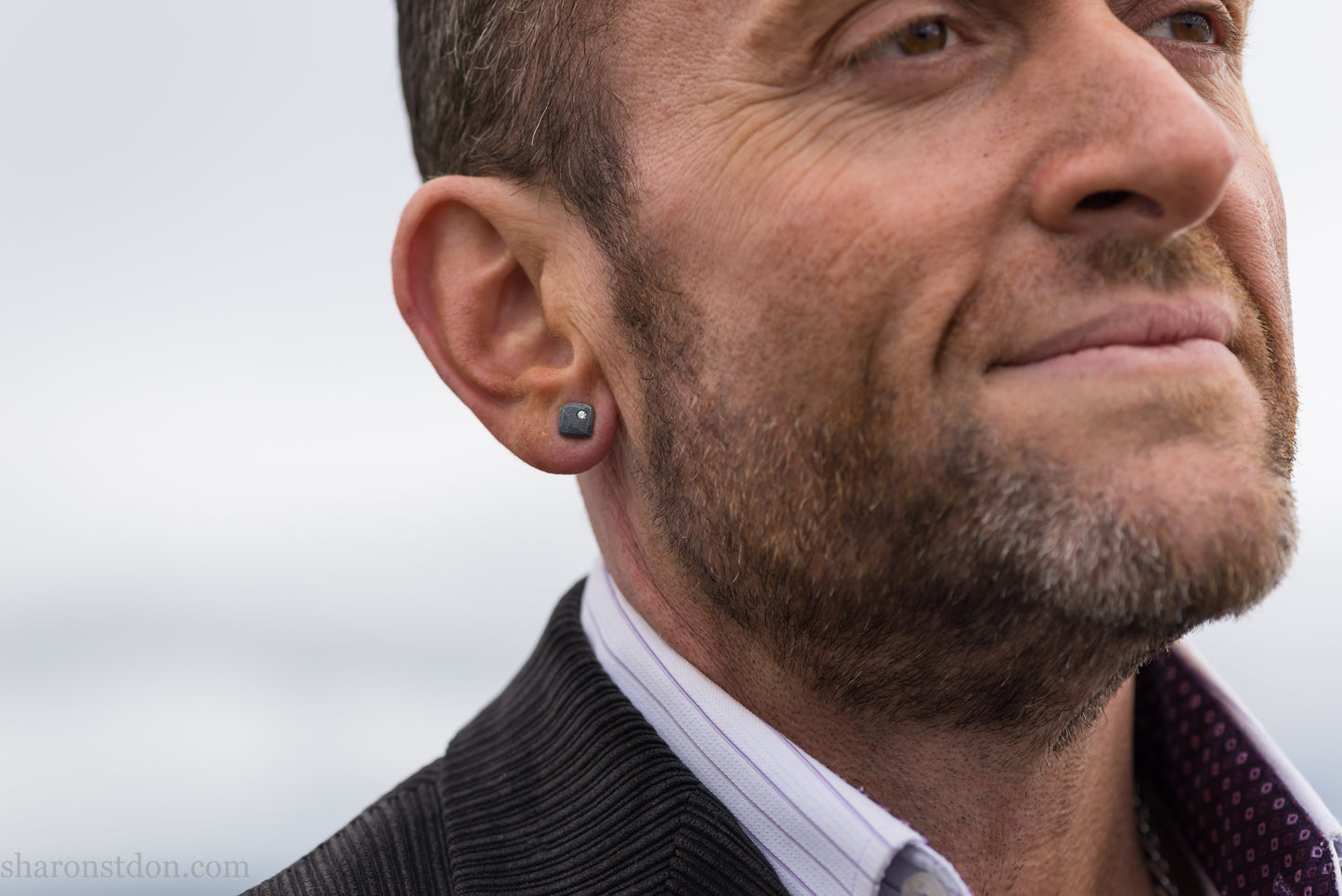 Man with beard wearing a handcrafted 7mm square sterling silver stud earring, oxidized black with a lab-grown diamond moissanite set in the corner.