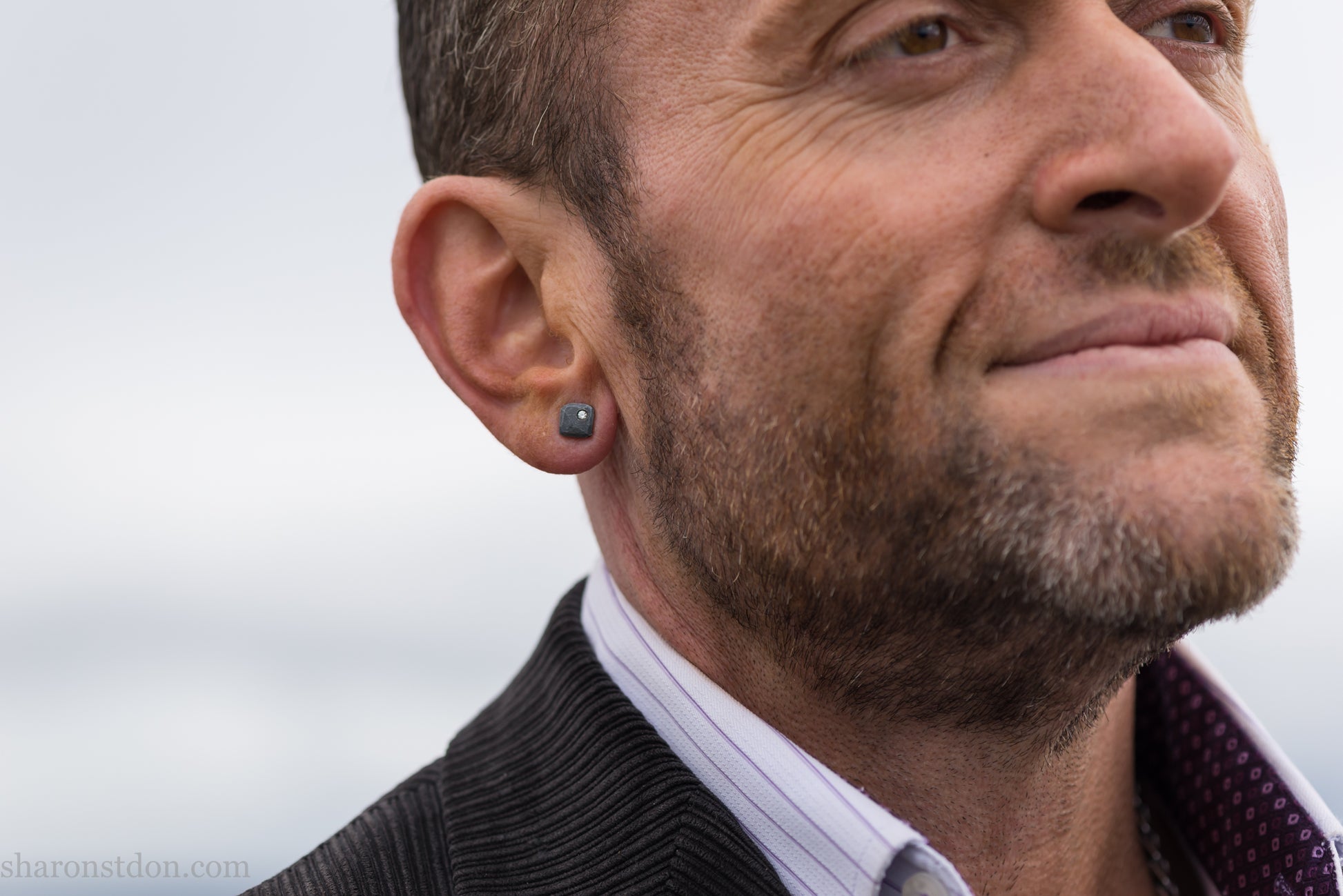 Man with beard wearing a handcrafted 7mm square sterling silver stud earring, oxidized black with a lab-grown diamond moissanite set in the corner.