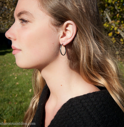 25mm oxidized black sterling silver round hammered hoop earring, close-up on model’s ear.