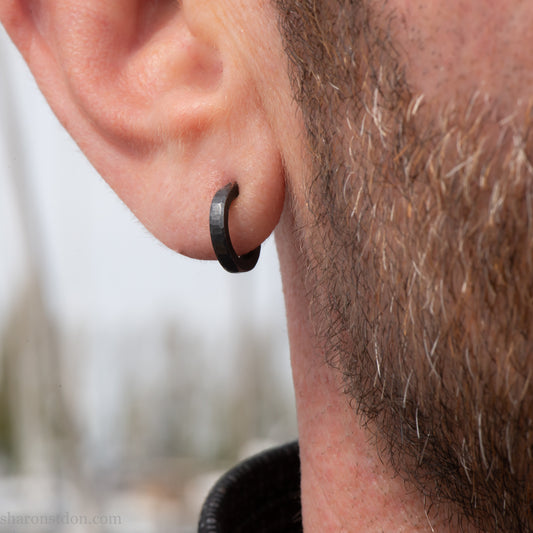 Solid 925 sterling silver hoop earrings for men with an oxidized matte black finish. 12mm exterior diameter, 9mm interior, 2mm wide, and 1.5mm thick. Solid sterling silver posts and backs. Handcrafted, each pair hand hammered by Sharon Saint Don in the Pacific Northwest of the USA. Worn by a man with light skin, dark hair and beard.