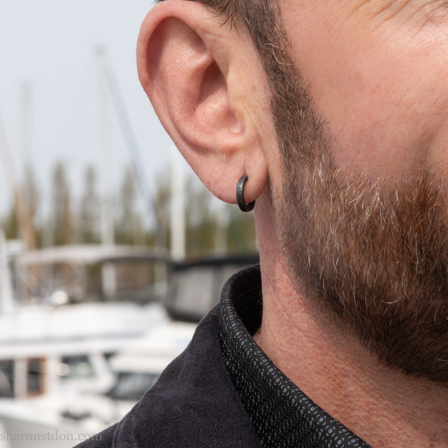 Solid 925 sterling silver hoop earrings for men with an oxidized matte black finish. 12mm exterior diameter, 9mm interior, 2mm wide, and 1.5mm thick. Solid sterling silver posts and backs. Handcrafted, each pair hand hammered by Sharon Saint Don in the Pacific Northwest of the USA. Worn by a man with light skin, dark hair and beard.