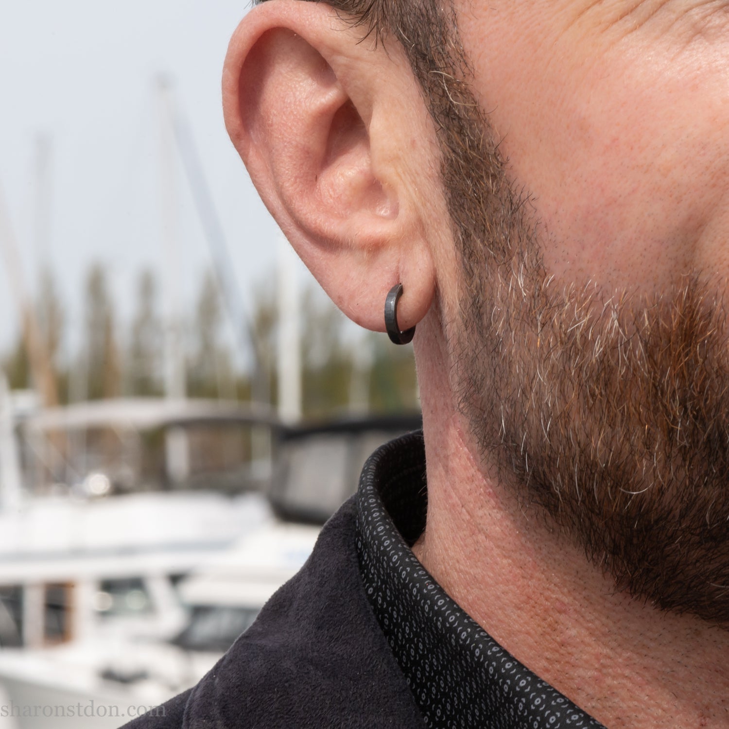 Solid 925 sterling silver hoop earrings for men with an oxidized matte black finish. 12mm exterior diameter, 9mm interior, 2mm wide, and 1.5mm thick. Solid sterling silver posts and backs. Handcrafted, each pair hand hammered by Sharon Saint Don in the Pacific Northwest of the USA. Worn by a man with light skin, dark hair and beard.
