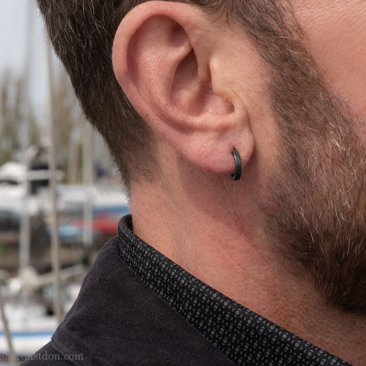 Solid 925 sterling silver hoop earrings for men with an oxidized matte black finish. 12mm exterior diameter, 9mm interior, 2mm wide, and 1.5mm thick. Solid sterling silver posts and backs. Handcrafted, each pair hand hammered by Sharon Saint Don in the Pacific Northwest of the USA. Worn by a man with light skin, dark hair and beard.