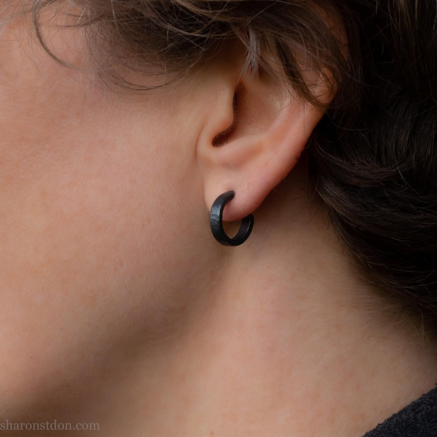 Close up photo of a person wearing oxidized black, handcrafted 14mm x 3mm hammered solid sterling silver hoop earrings.