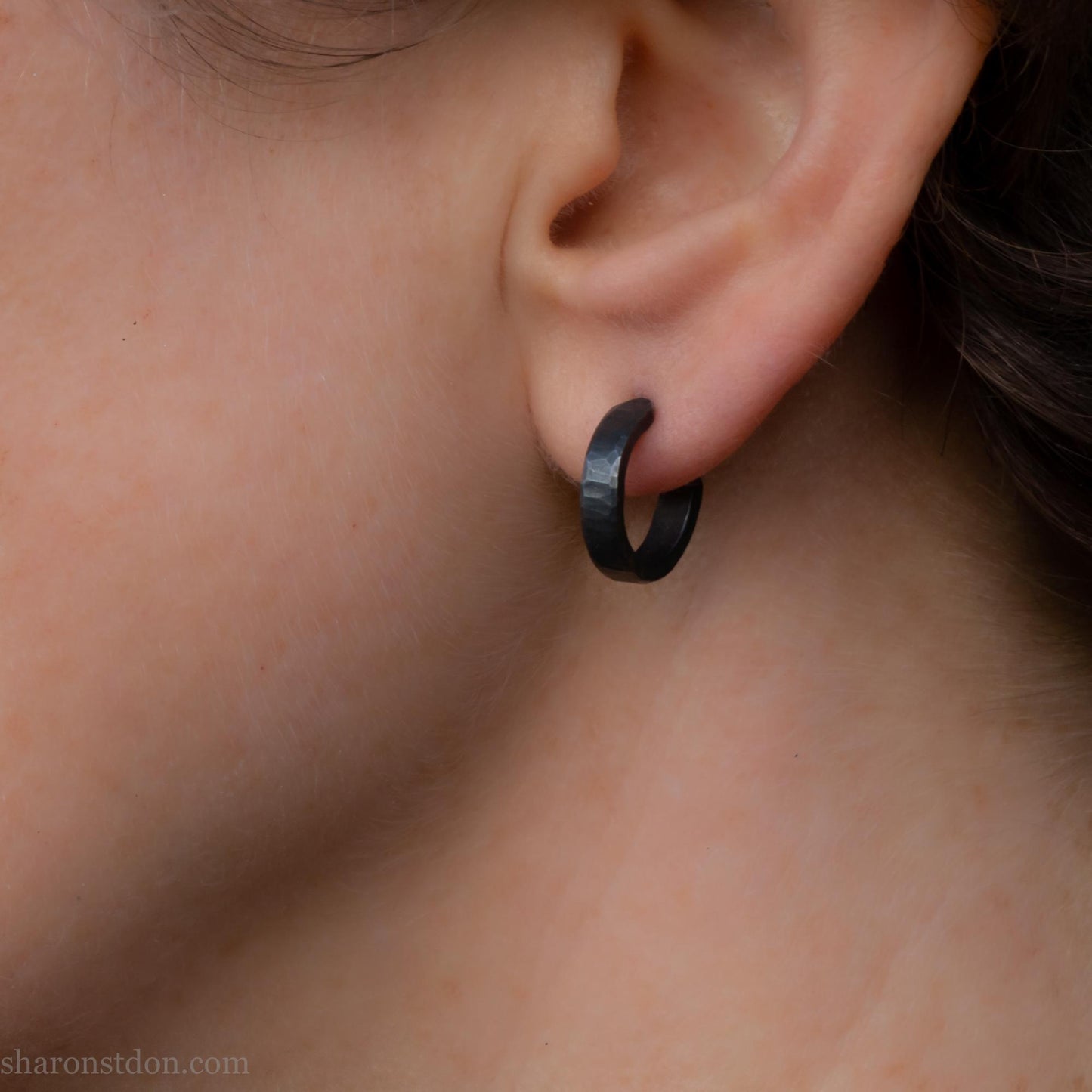 Close up photo of a person wearing oxidized black, handcrafted 14mm x 3mm hammered solid sterling silver hoop earrings.