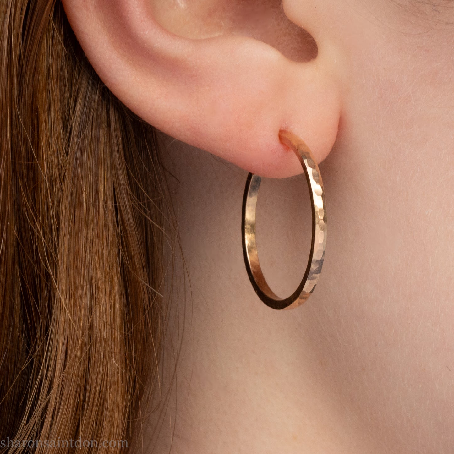 Solid hammered yellow gold hoop earrings with brushed, matte finish, gold posts and  locking backs; worn on ear.