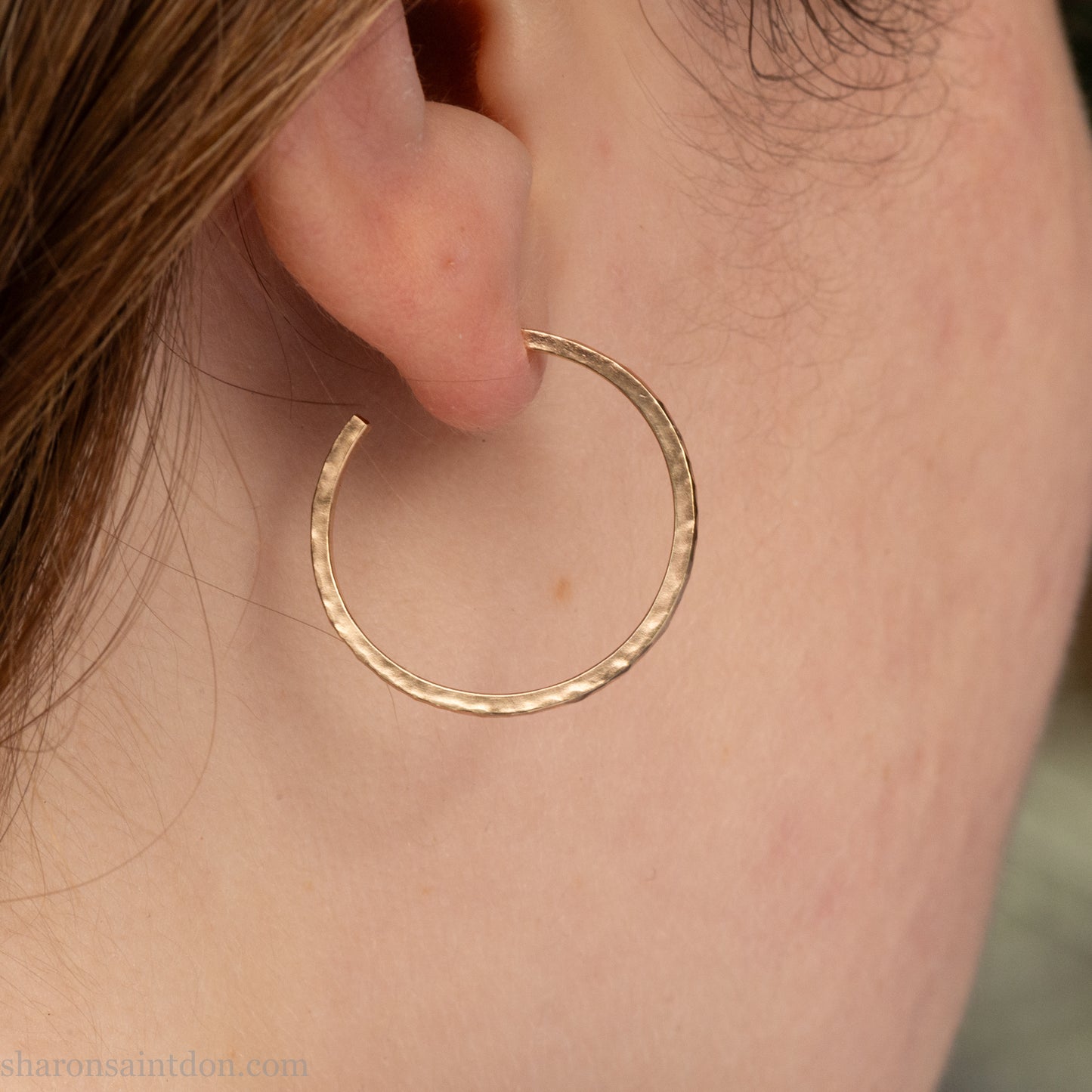 Solid hammered yellow gold hoop earrings with brushed, matte finish, gold posts and  locking backs; worn on ear.