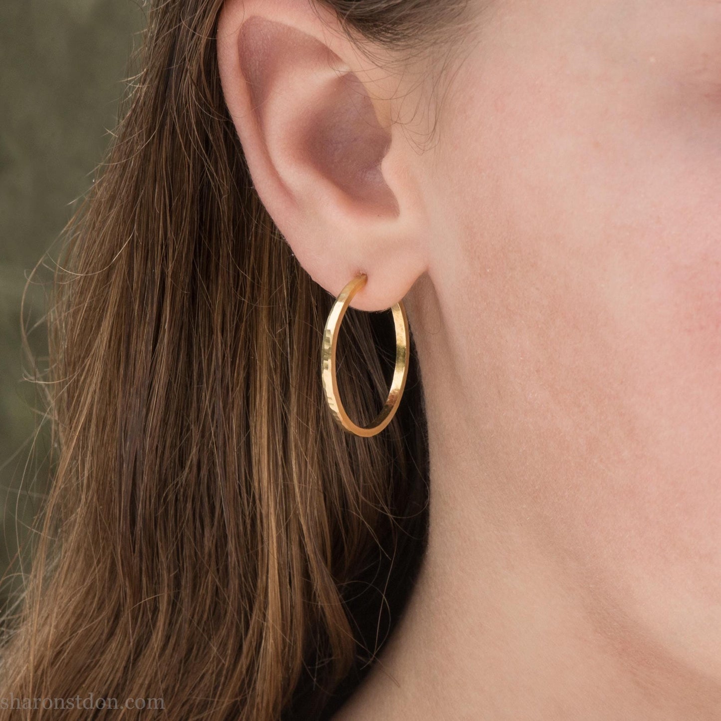 Solid 14k gold hoop earrings handmade in North America by Sharon SaintDon. Solid hammered yellow gold with a matte brushed finish. 25mm diameter round, 1.5mm thick, solid gold posts and backs. Worn by a teen girl with light skin and brown hair.