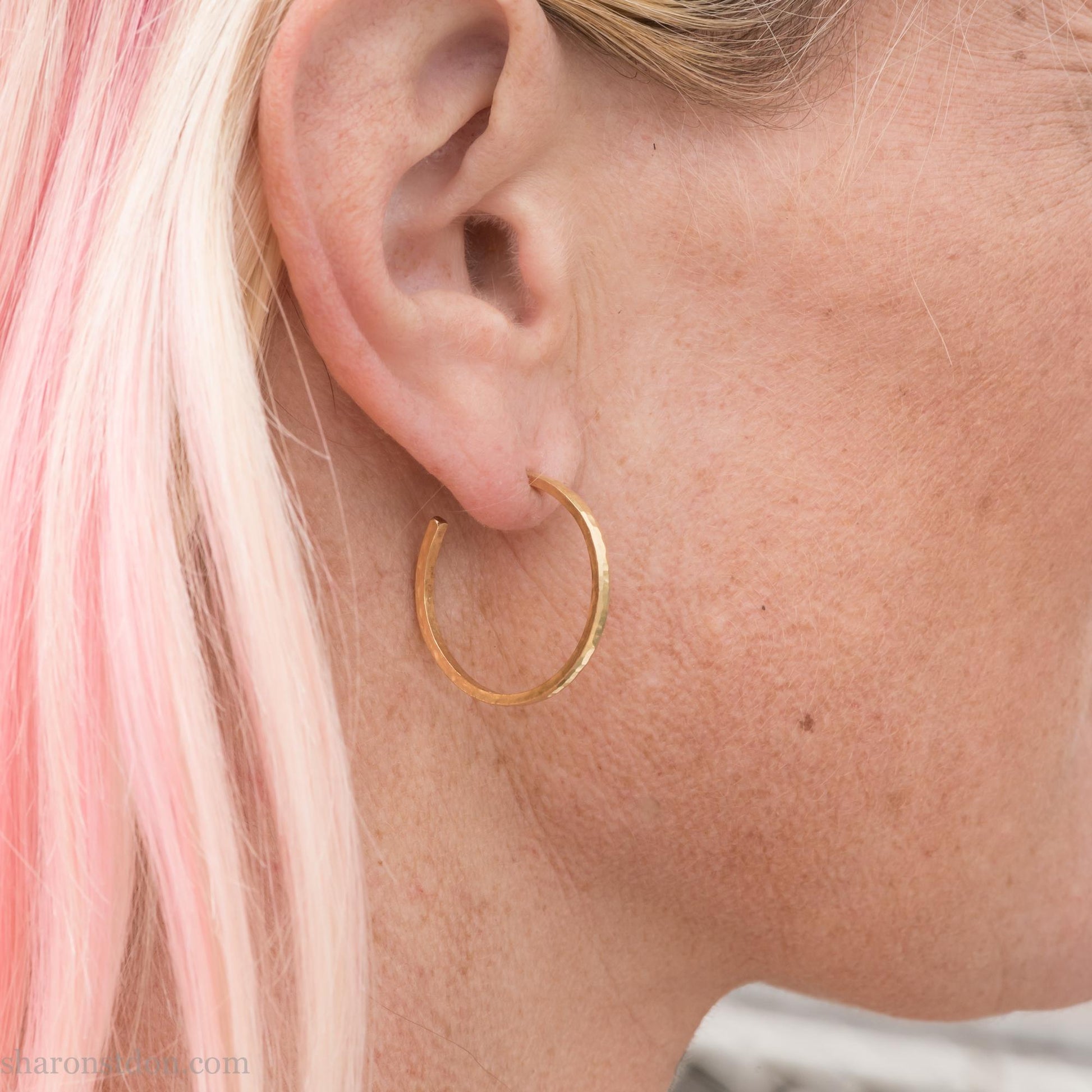 Solid 14k gold hoop earrings handmade in North America by Sharon SaintDon. Solid hammered yellow gold with a matte brushed finish. 25mm diameter round, 1.5mm thick, solid gold posts and backs. Worn by a woman with light skin and blonde hair.