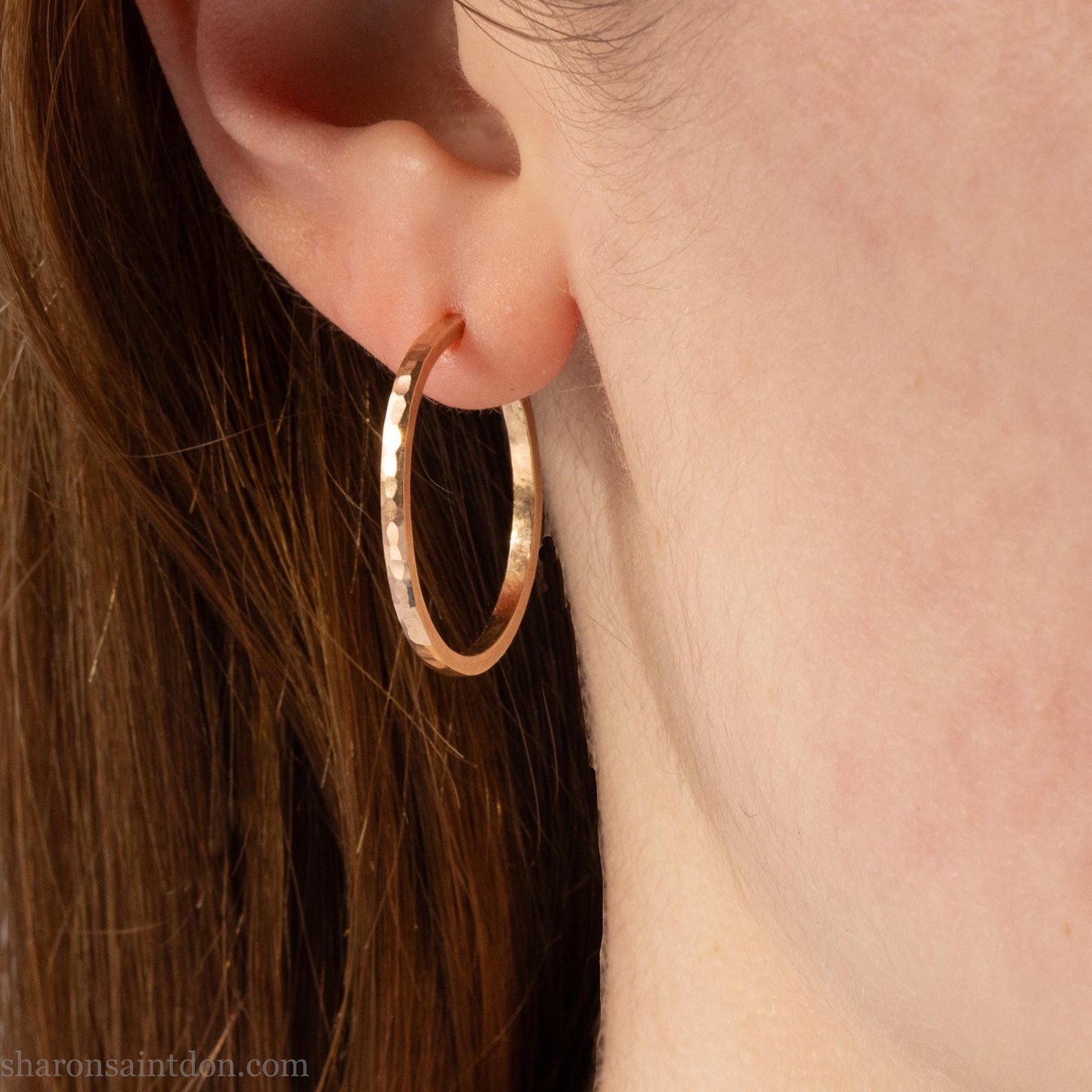 Solid 14k gold hoop earrings handmade in North America by Sharon SaintDon. Solid hammered yellow gold with a matte brushed finish. 25mm diameter round, 1.5mm thick, solid gold posts and backs. Worn by a young woman with light skin and brown hair.