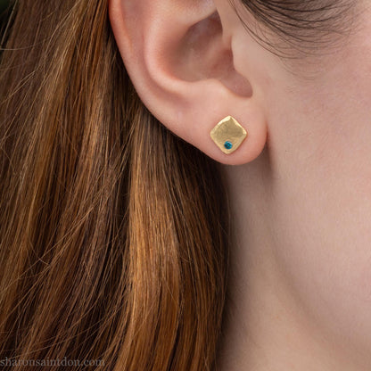 Solid yellow gold square stud earrings with 2mm blue diamonds, matte finish, shown worn on a model (side view, one ear).