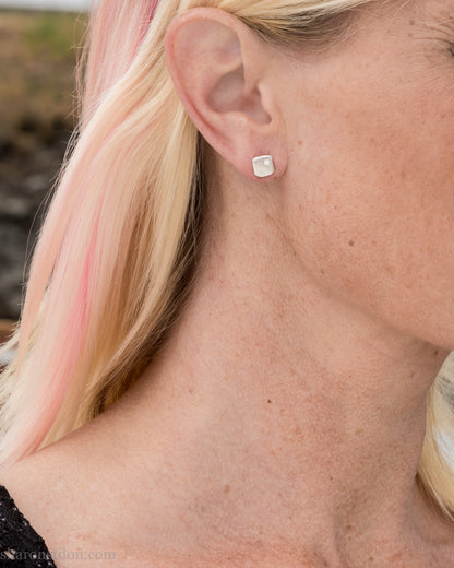 Solid 925 sterling silver stud earrings with lab-grown moissanite, 7mm square, hammered silver with brushed matte finish, corner-set stone, sterling silver posts and backs. Worn by a model with blonde hair.