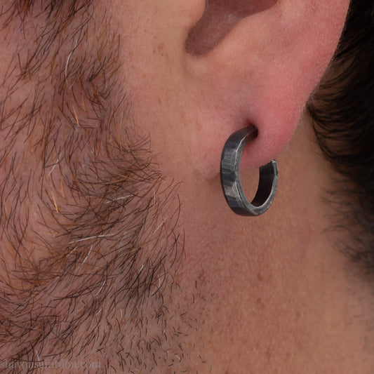 Solid 925 sterling silver hoop earrings for men with a shiny, matte finish. 18mm Solid 925 sterling silver hoop earrings with an oxidized, matte, black finish. 18mm exterior diameter, 15mm interior, 3mm wide, and 1.5mm thick. Solid sterling silver posts and backs. Handcrafted in the USA, each pair hand hammered by Sharon Saint Don. Worn by a man with light skin, dark hair and beard.