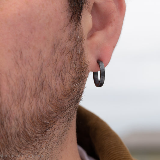 Solid 925 sterling silver hoop earrings with an oxidized, matte, black finish. 18mm exterior diameter, 15mm interior, 3mm wide, and 1.5mm thick. Solid sterling silver posts and backs. Handcrafted in the USA, each pair hand hammered by Sharon Saint Don. Worn by a man with light skin, dark hair and beard.