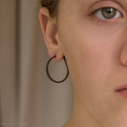 25mm oxidized black sterling silver round hammered hoop earring, close-up on model’s ear.