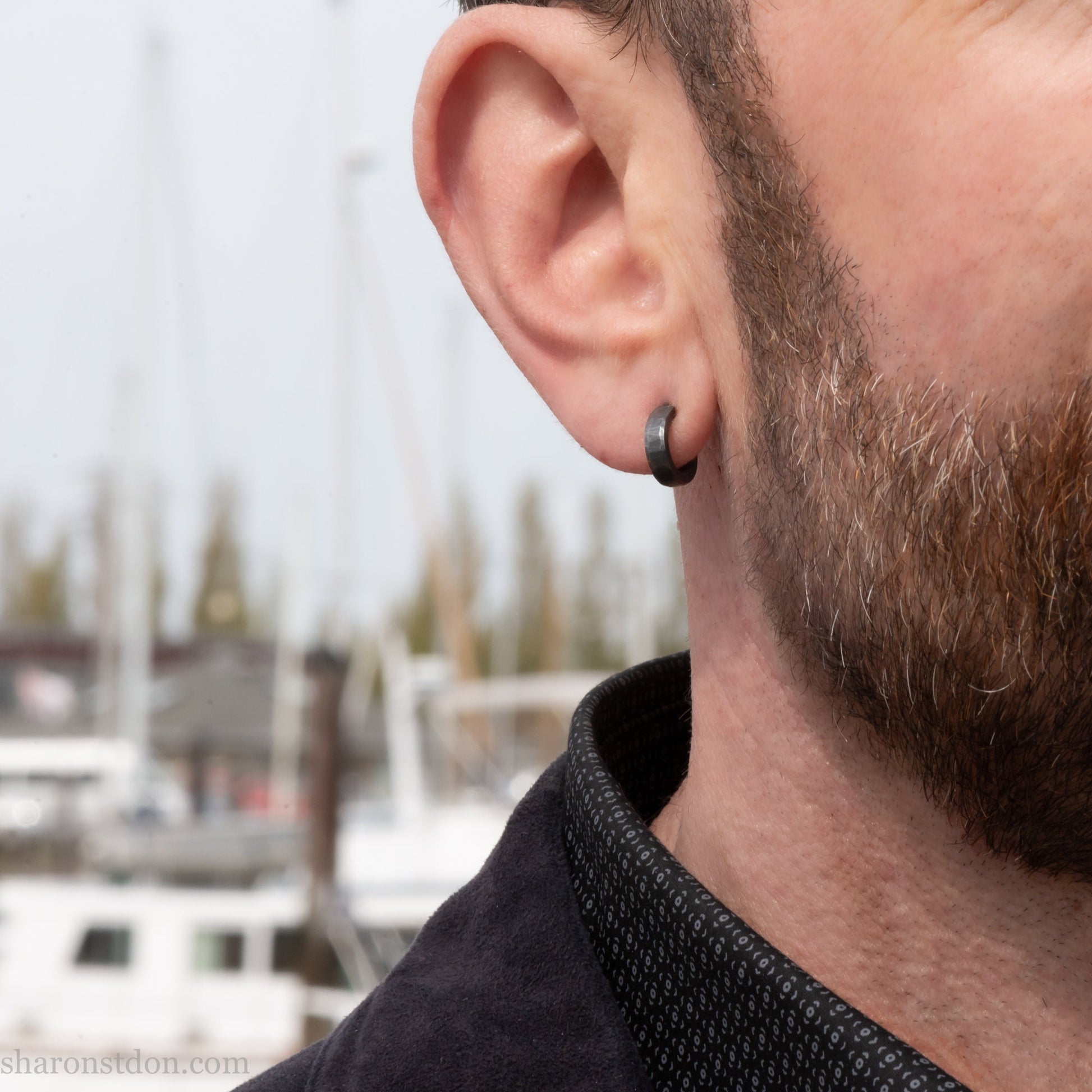Hammered solid 925 silver hoop earrings with an oxidized black finish. 12mm exterior diameter, 9mm interior, 3mm wide. and 1.5mm thick. Solid sterling silver posts and backs. Handcrafted by Sharon Saint Don in the USA. Pictured on a man with fair skin, medium brown hair and beard.