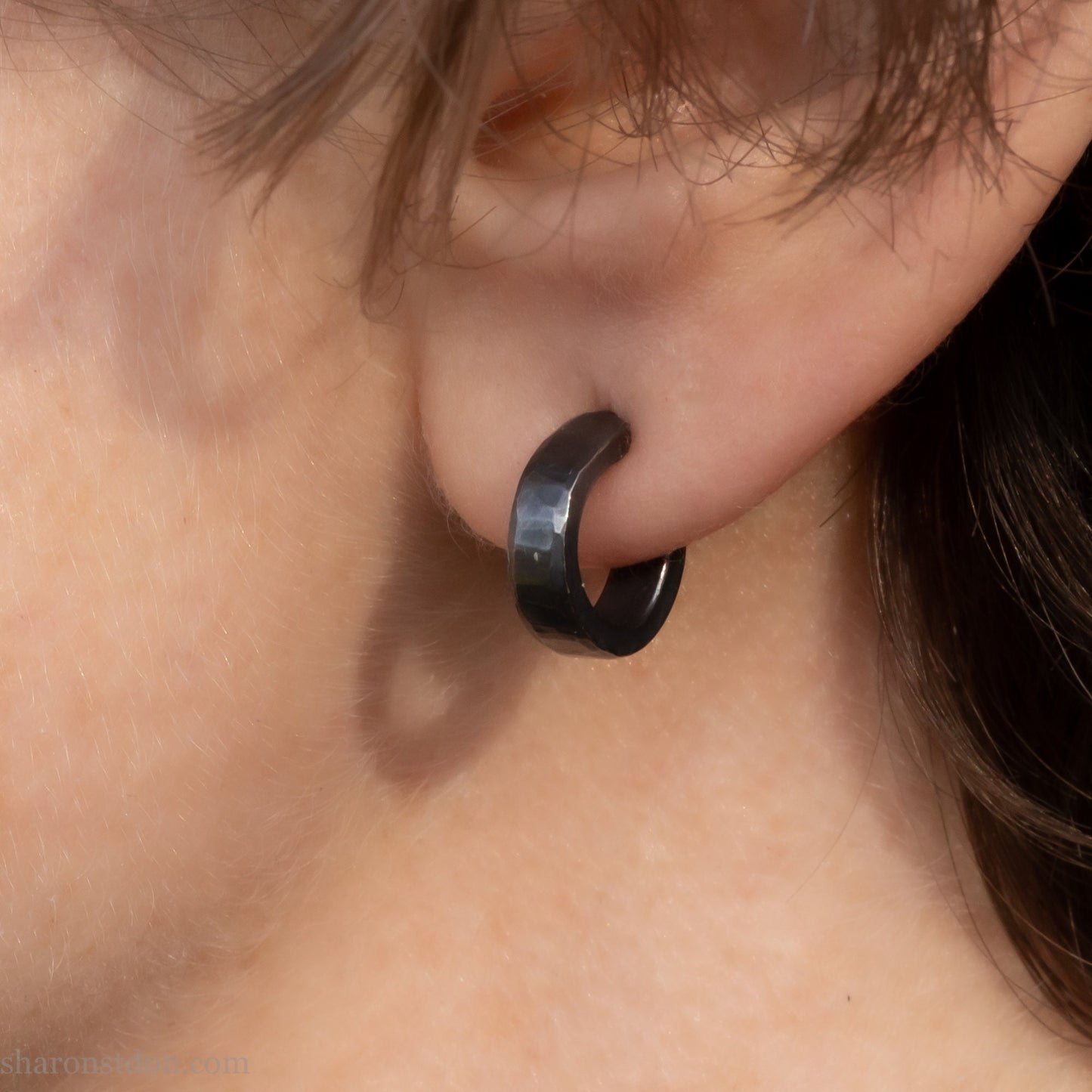 Hammered solid 925 silver hoop earrings with an oxidized black finish. 12mm exterior diameter, 9mm interior, 3mm wide. and 1.5mm thick. Solid sterling silver posts and backs. Handcrafted by Sharon Saint Don in the USA. Pictured on a person with fair skin and medium brown hair.