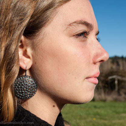38mm solid 925 sterling silver sculptural round disc earrings worn on ear, hand-punched pattern, flat matte black oxidized finish.
