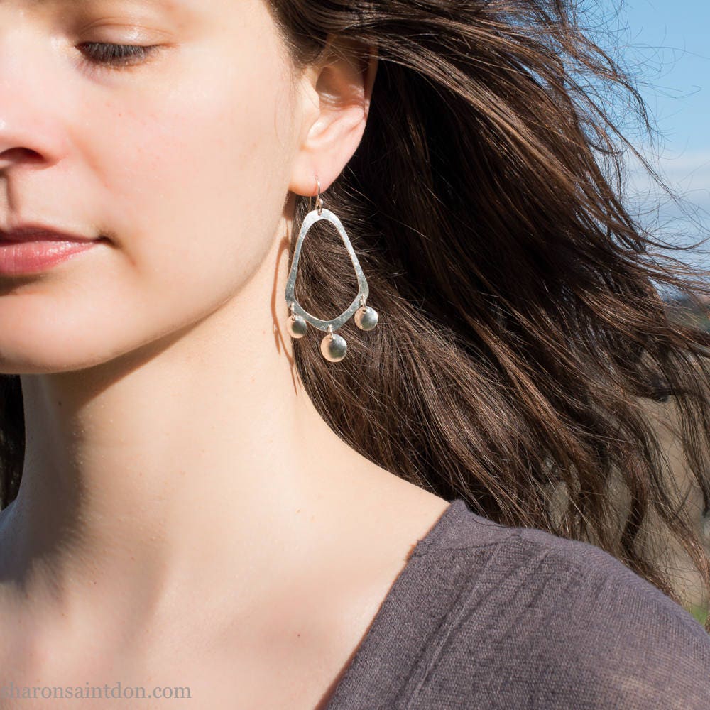 925 sterling silver handcrafted earrings. A hammered, angular, teardrop loop with three dots hanging from the angled teardrop points. Size: 60mm high to top of hook x 35mm at widest point, dots approximately 1cm. Comfortable, lightweight, with handmade ear wire hooks. Worn by a young woman with fair skin and dark brown hair.