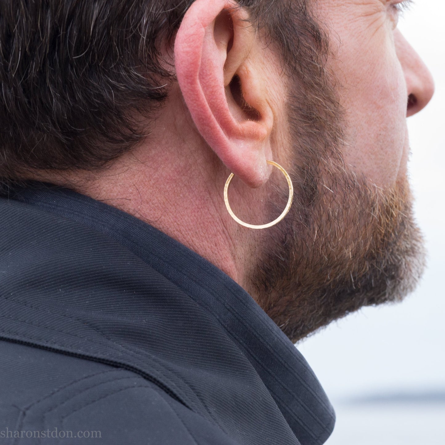 25mm diameter by 1.5mm thick handmade solid 14k yellow gold hoop earrings with solid 14k gold posts and backs. Handmade by Sharon SaintDon in North America. Hammered from solid gold with a matte finish. Worn by a man with light skin, dark beard and hair.