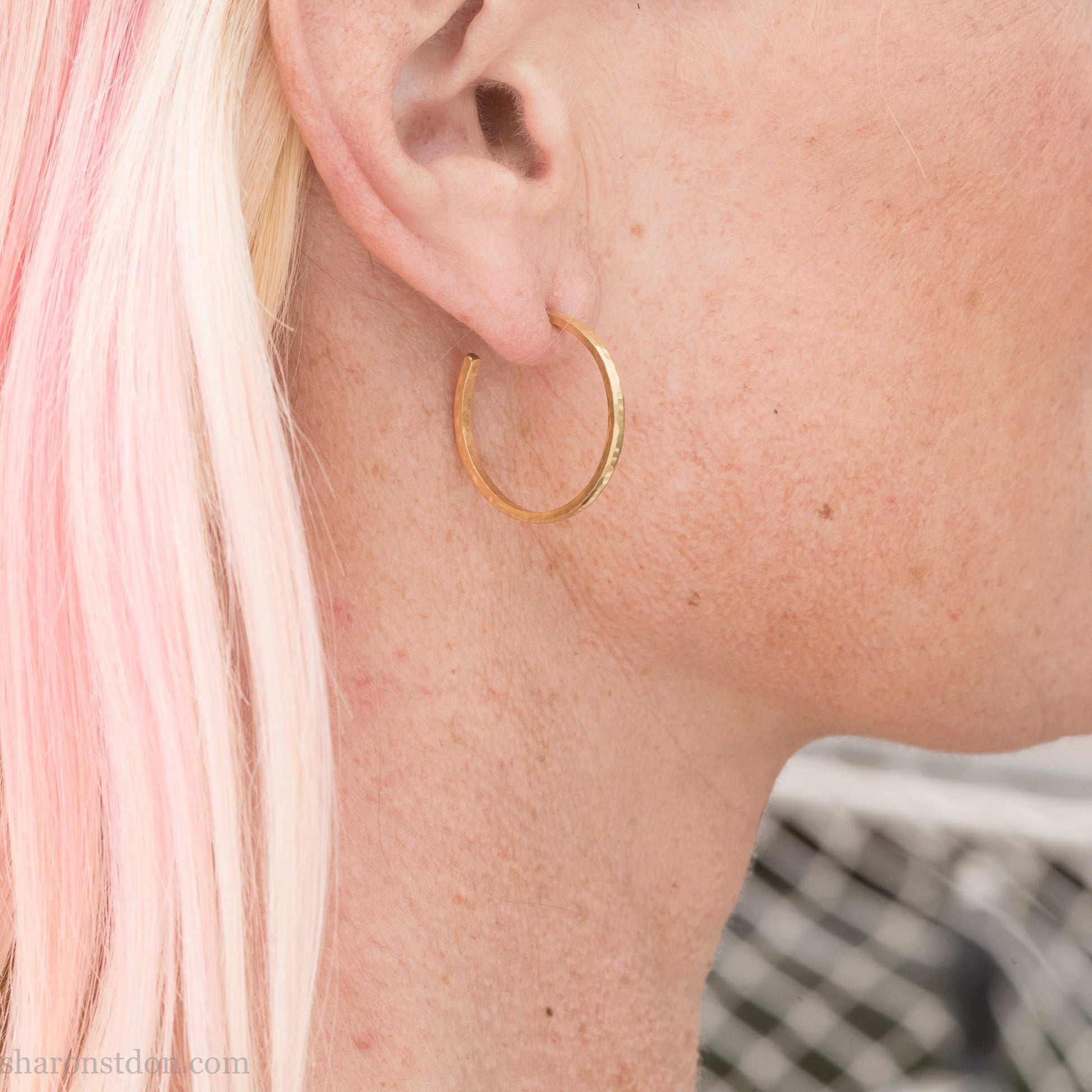 25mm diameter by 1.5mm thick handmade solid 14k yellow gold hoop earrings with solid 14k gold posts and backs. Handmade by Sharon SaintDon in North America. Hammered from solid gold with a matte finish. Worn by a woman with light skin and blonde hair.