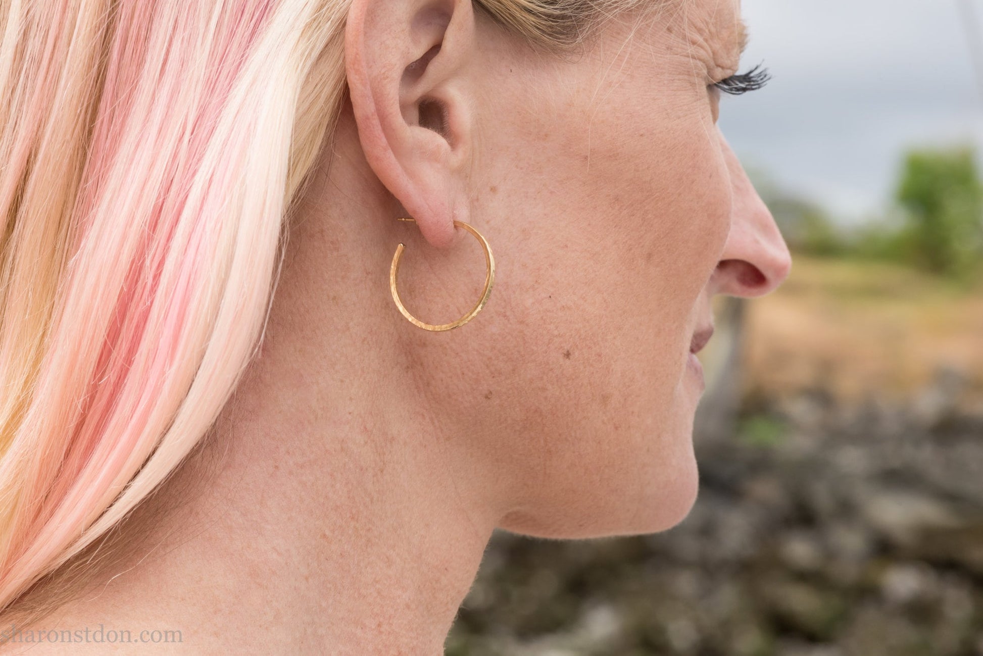 25mm diameter by 1.5mm thick handmade solid 14k yellow gold hoop earrings with solid 14k gold posts and backs. Handmade by Sharon SaintDon in North America. Hammered from solid gold with a matte finish. Worn by a woman with light skin and blonde hair.