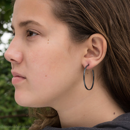 Black 925 sterling silver hoop earrings, 40mm diameter, 2mm wide, 1mm thick. Hammered solid silver with silver posts and backs, worn by a person.