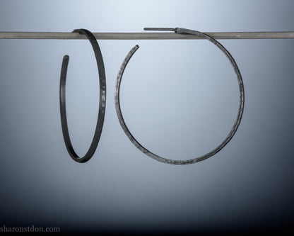 Black 925 sterling silver hoop earrings, 40mm diameter, 2mm wide, 1mm thick. Hammered solid silver with silver posts and backs, worn by a person.