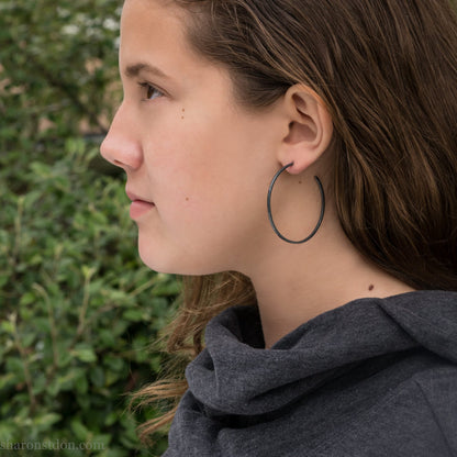 Black 925 sterling silver hoop earrings, 40mm diameter, 2mm wide, 1mm thick. Hammered solid silver with silver posts and backs, worn by a person.