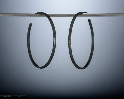 Black 925 sterling silver hoop earrings, 40mm diameter, 2mm wide, 1mm thick. Hammered solid silver with silver posts and backs, worn by a person.