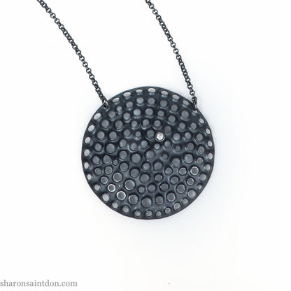 Large round, handcrafted sterling silver pendant necklace with perforated holes and oxidized black finish, accented with cubic zirconia, on black chain.