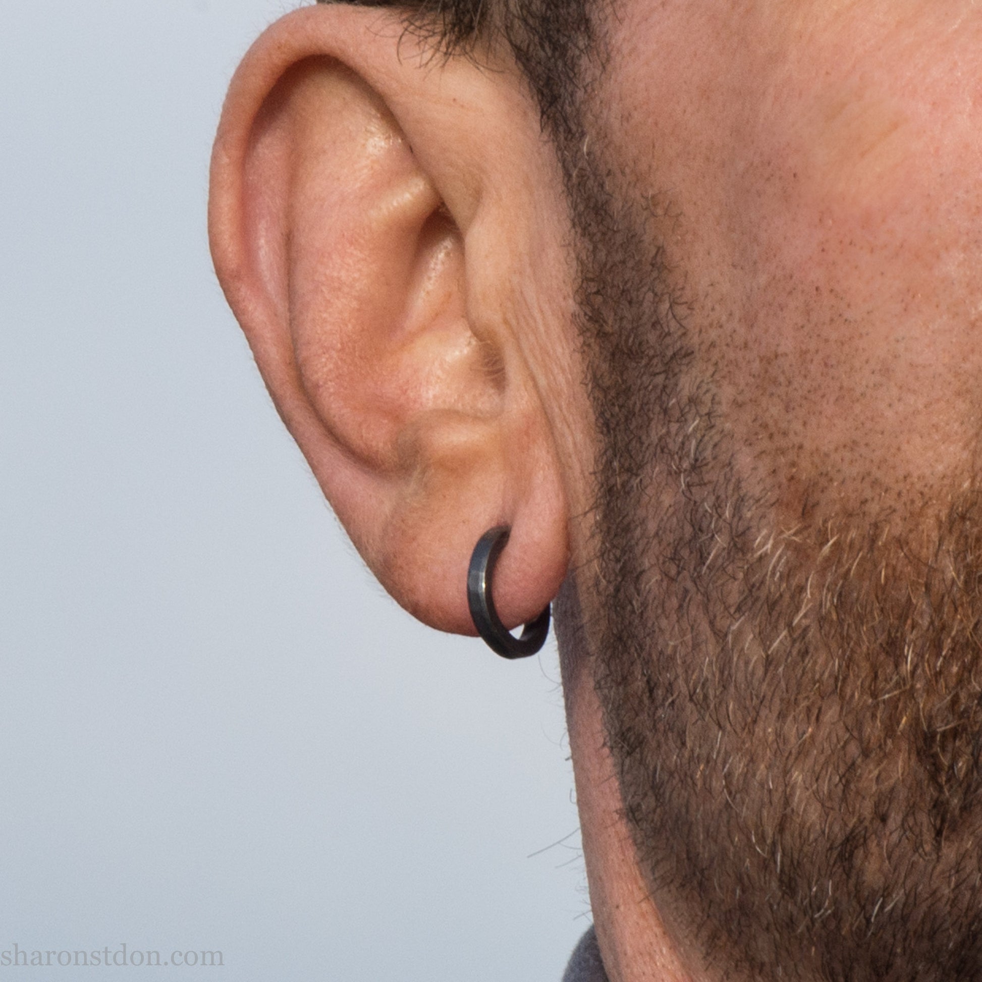 Hand hammered solid 925 sterling silver hoop earrings, oxidized black, 14mm diameter x 2mm wide, worn on an ear.