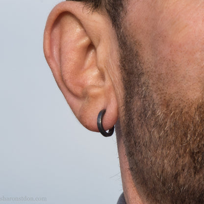 Hand hammered solid 925 sterling silver hoop earrings, oxidized black, 14mm diameter x 2mm wide, worn on an ear.