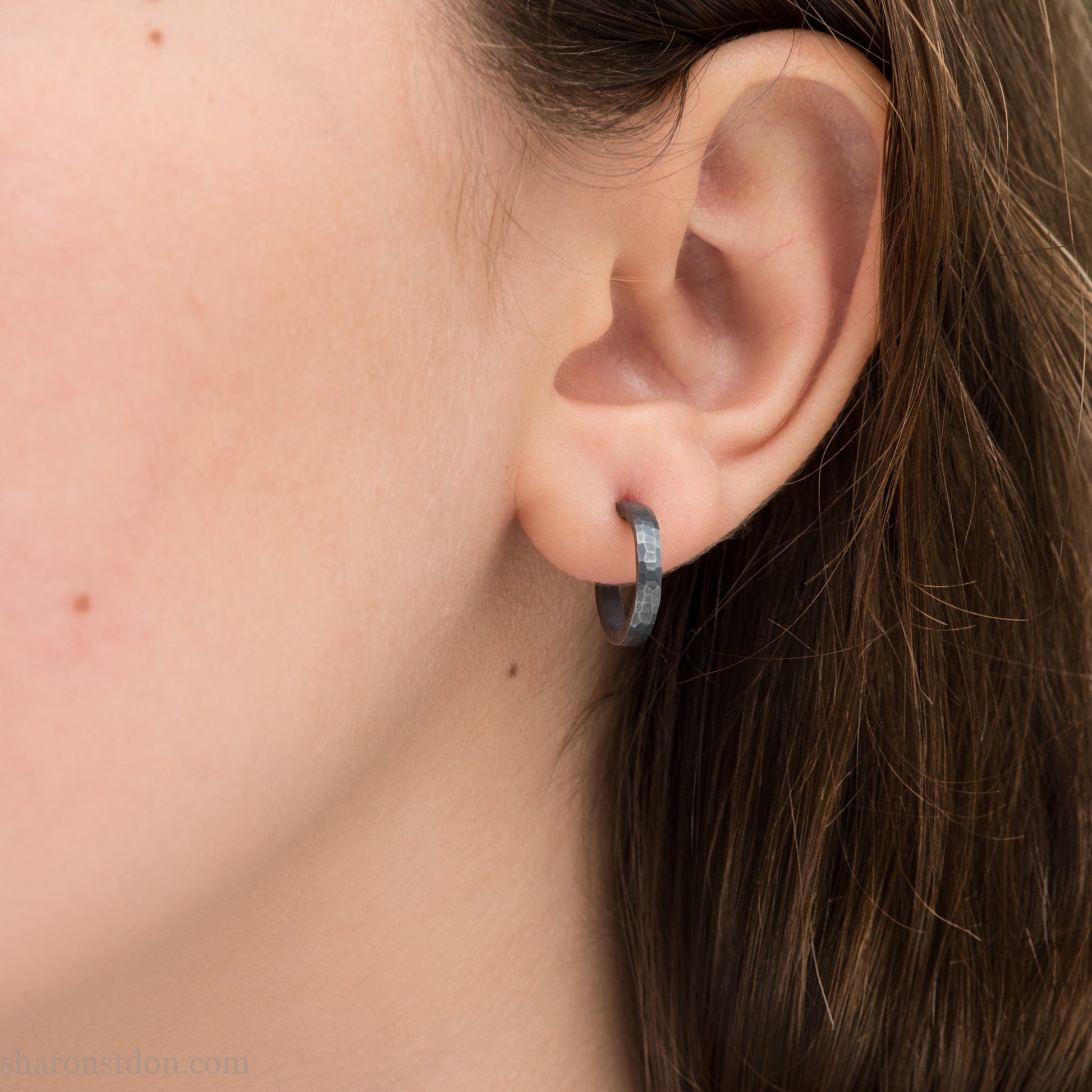 Hand hammered solid 925 sterling silver hoop earrings, oxidized black, 14mm diameter x 2mm wide, worn on an ear.