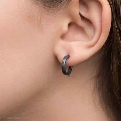 Hand hammered solid 925 sterling silver hoop earrings, oxidized black, 14mm diameter x 2mm wide, worn on an ear.