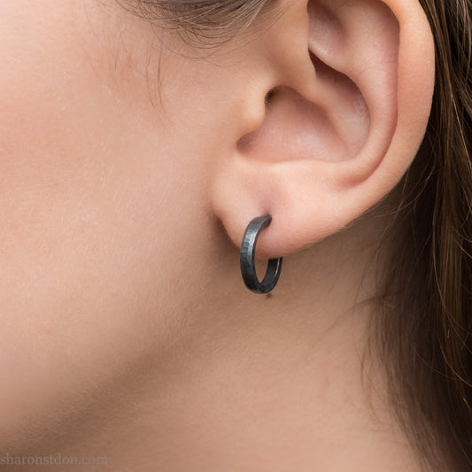 Hand hammered solid 925 sterling silver hoop earrings, oxidized black, 14mm diameter x 2mm wide, worn on an ear.