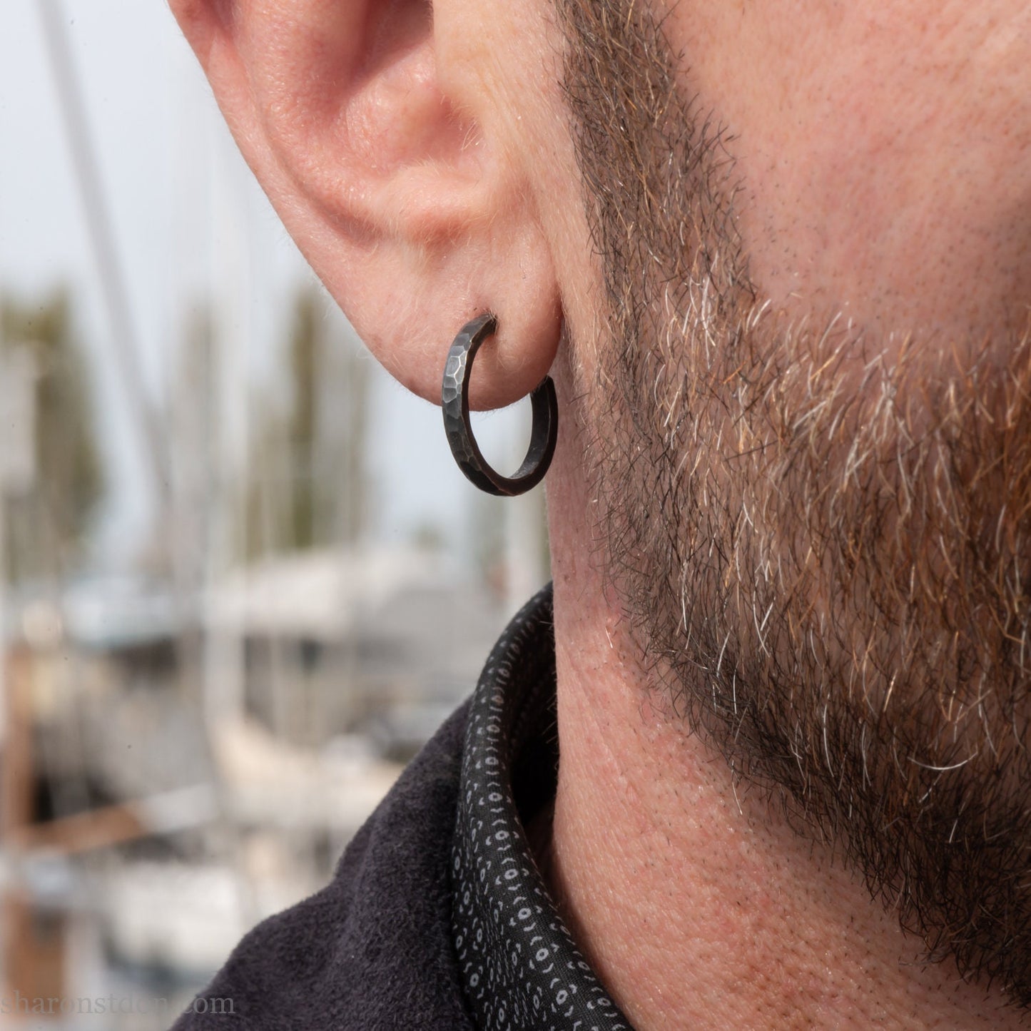 A close-up of a man's ear wearing a 18x2mm matte dark patina 925 sterling silver hammered hoop earring, with a blurred outdoor marina background.