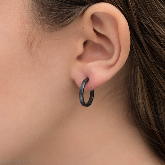 Oxidized black sterling silver 18x2mm hammered hoop earrings worn on ear by woman, side profile.