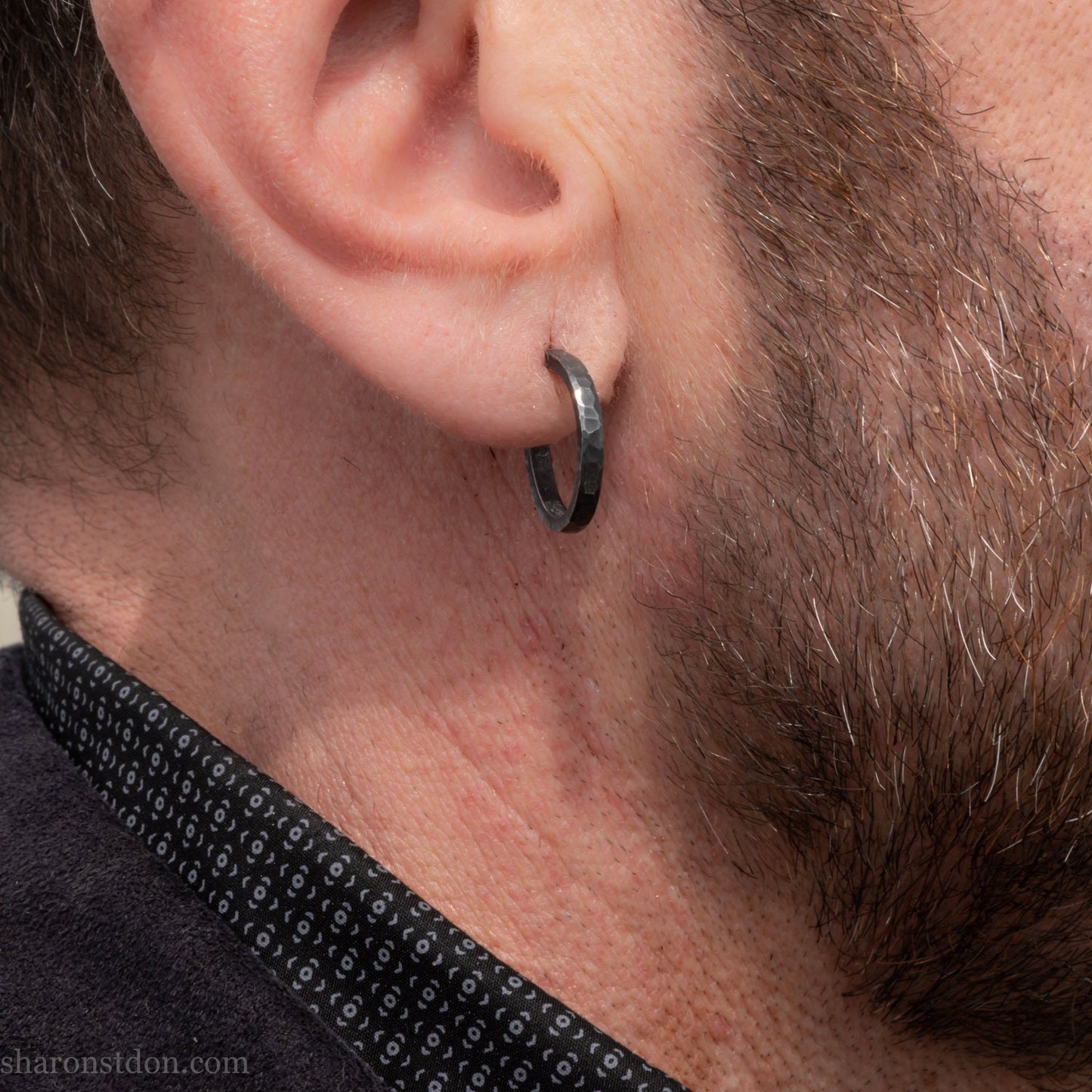 A close-up of a man's ear wearing a 18x2mm matte dark patina 925 sterling silver hammered hoop earring, with a blurred outdoor marina background.