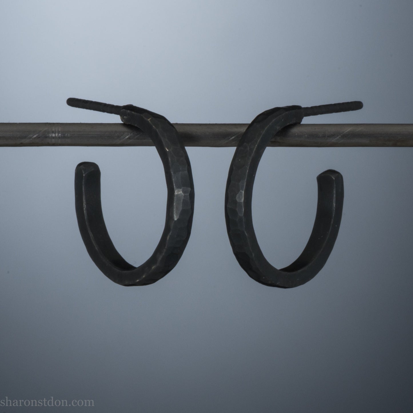18x2mm oxidized black 925 sterling silver hammered hoop earrings with a matte dark patina, shown on display bar.