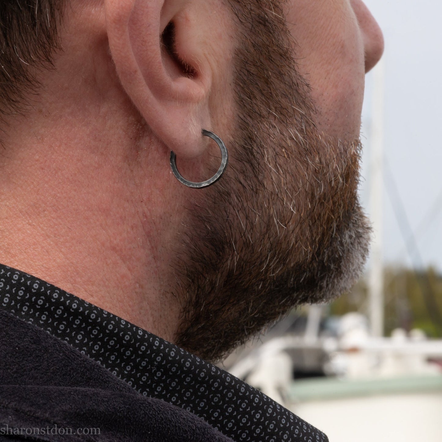 A close-up of a man's ear wearing a 18x2mm matte dark patina 925 sterling silver hammered hoop earring, with a blurred outdoor marina background.