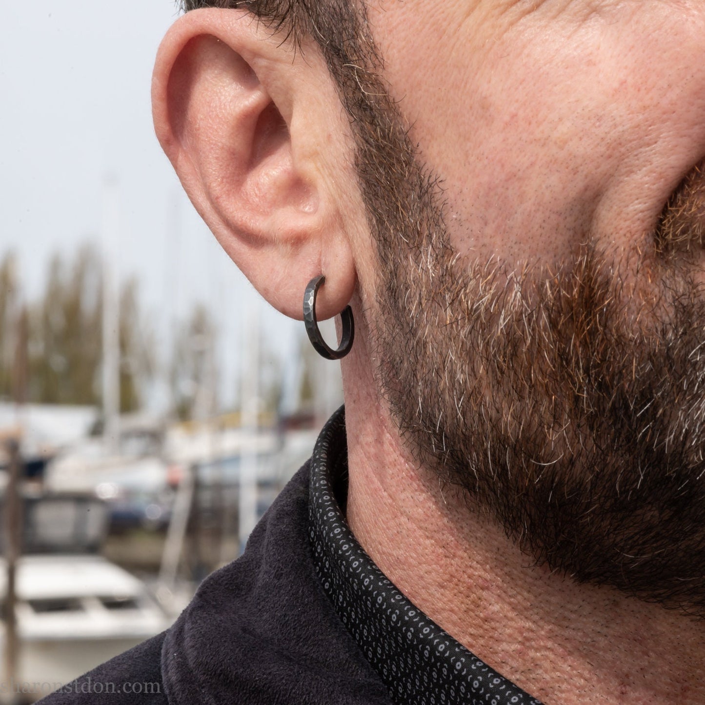 A close-up of a man's ear wearing a 18x2mm matte dark patina 925 sterling silver hammered hoop earring, with a blurred outdoor marina background.