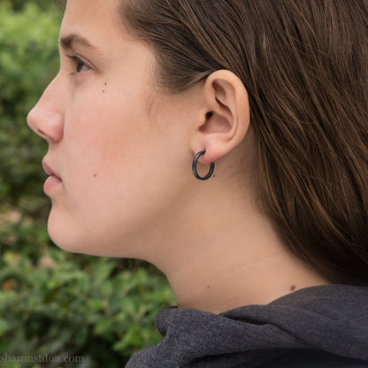 Oxidized black sterling silver 18x2mm hammered hoop earrings worn on ear by woman, side profile.
