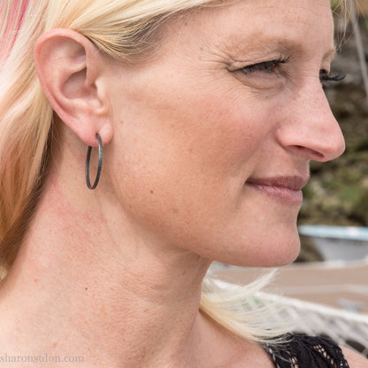 Oxidized black sterling silver hammered round hoop earring, shown worn on a model’s ear. 30mm diameter, 2mm wide, solid hammered silver.