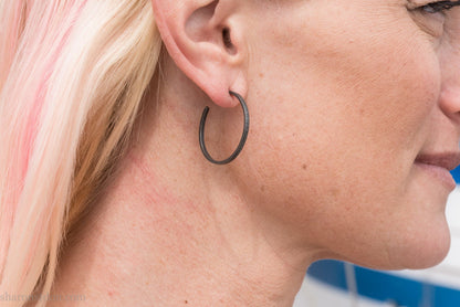 Oxidized black sterling silver hammered round hoop earring, shown worn on a model’s ear. 30mm diameter, 1.5mm wide, solid hammered silver.